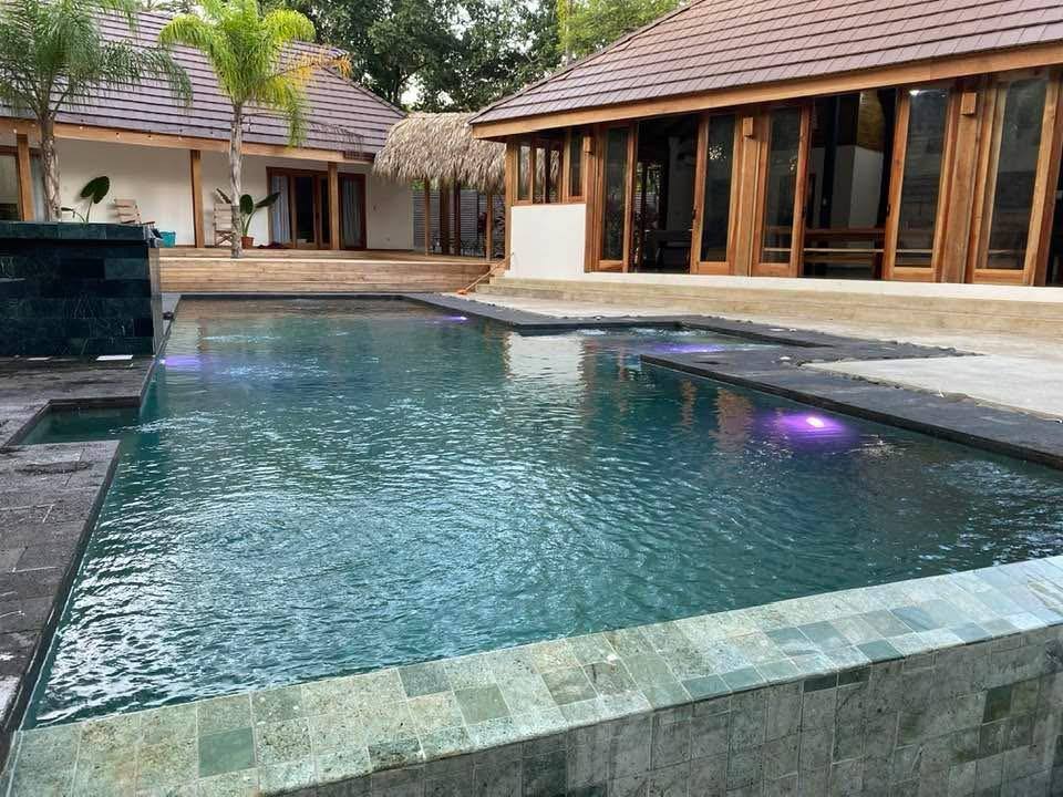 Piscina terminada con acabado de piedra natural Sukabumi e iluminación, frente a una casa con techo de teja